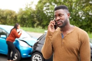 African-American man talks on cellphone while driver takes pictures of vehicle damage.